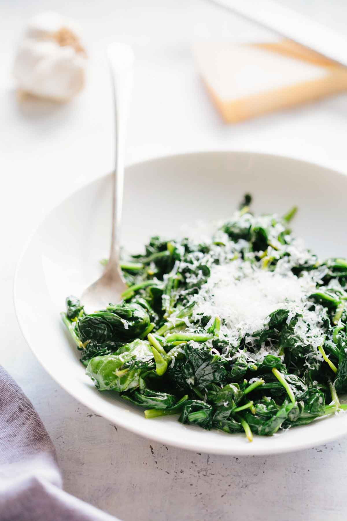 Another angle of sautéed spinach topped with grated parmesan cheese in a white bowl with a spoon, showing more detail of the bright green leaves and cheese.