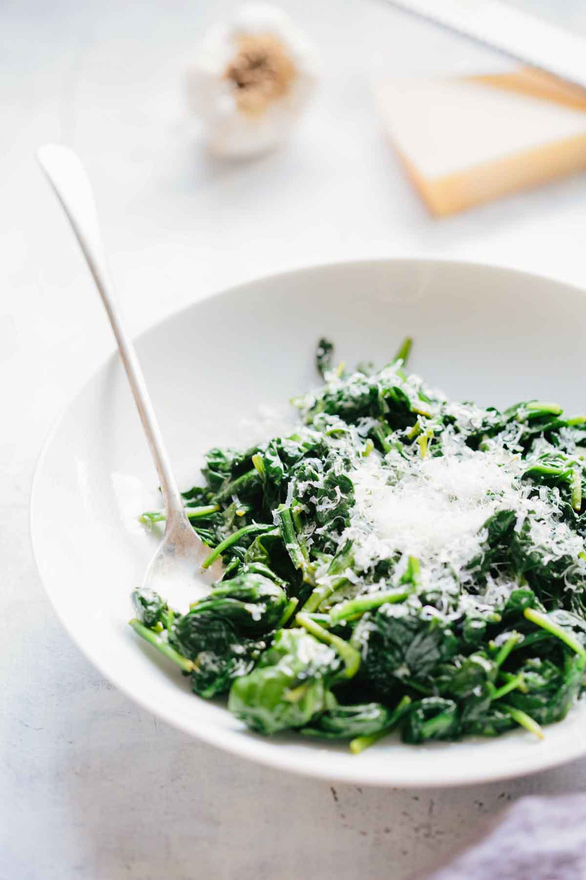Close-up of sautéed spinach with parmesan cheese in a white bowl with a silver spoon, garlic and cheese visible blurred in the background.