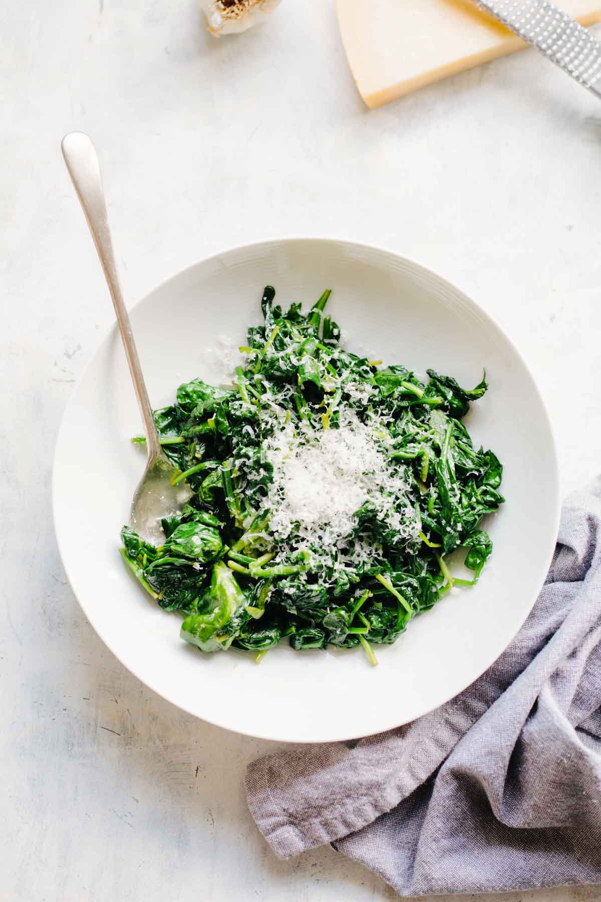 Overhead view of sautéed spinach with parmesan in a white bowl with a silver spoon on a white marble surface with a gray linen napkin and cheese visible in the corner.