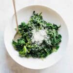 Directly overhead view of a white bowl filled with bright green sautéed spinach topped with freshly grated parmesan cheese and a silver spoon against a white surface.