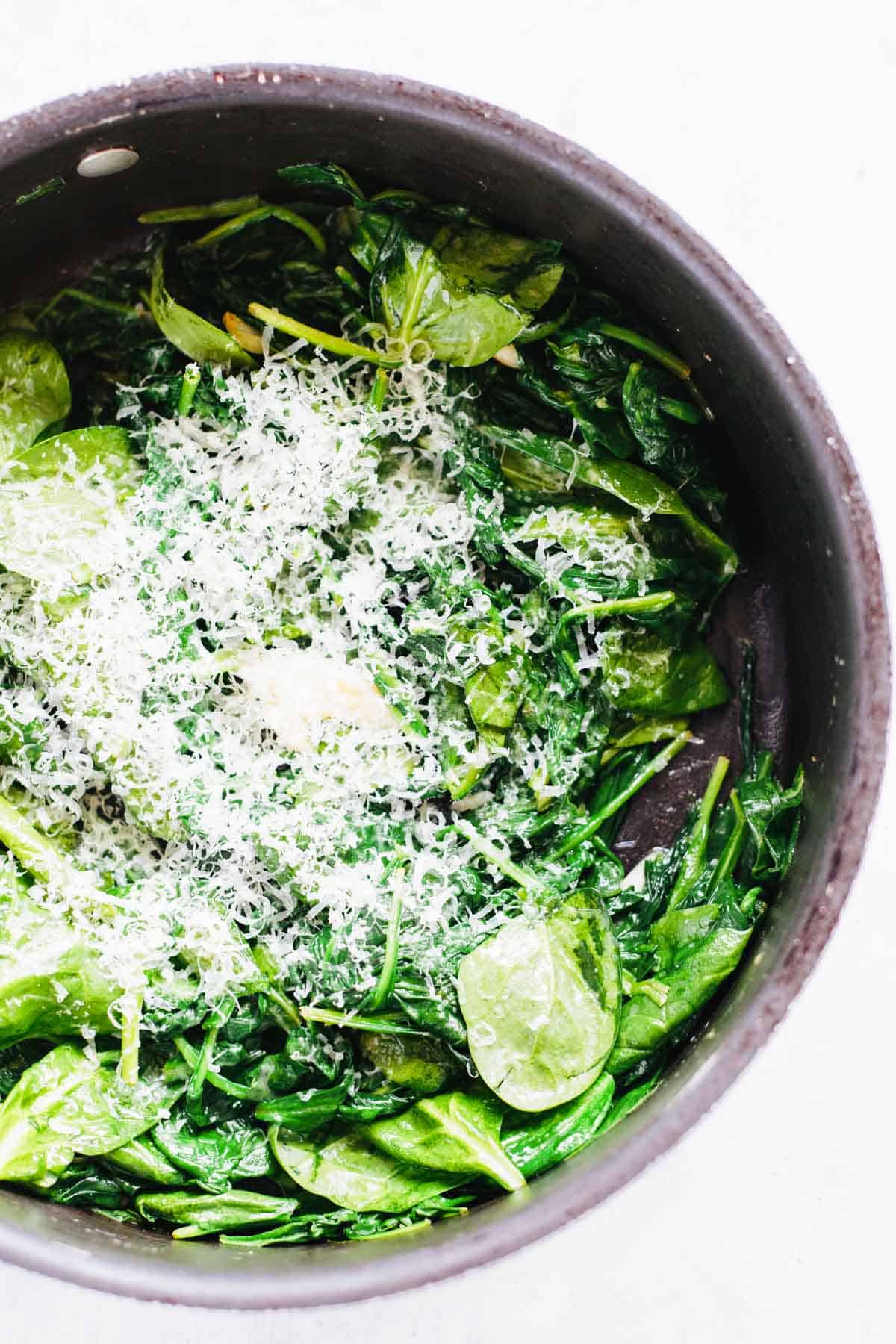 Top-down view of sautéed spinach with freshly grated parmesan cheese in a dark pan or skillet, showing the bright green leaves and white cheese.