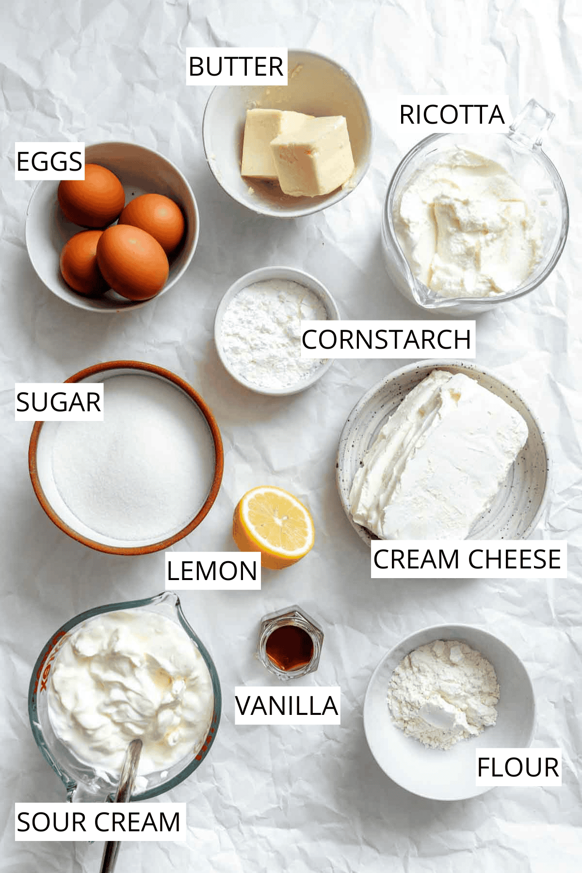 Overhead view of ingredients for ricotta cheesecake on white parchment paper, with labeled components including eggs, butter, ricotta, cornstarch, cream cheese, flour, sugar, lemon, vanilla, and sour cream.