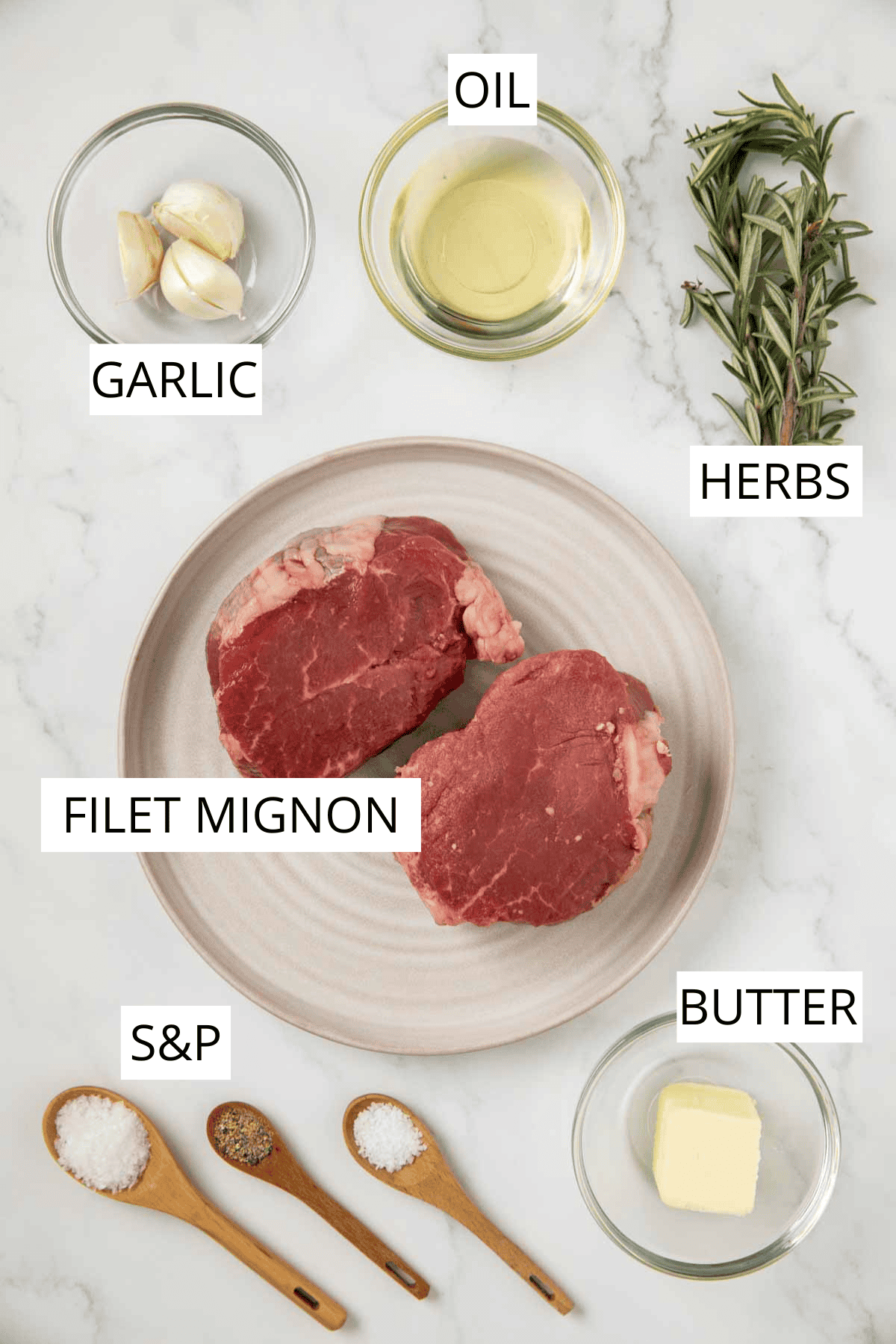 A top-down view of recipe ingredients arranged on a white marble countertop. Two raw filet mignon steaks on a beige plate surrounded by small bowls containing garlic cloves, oil, butter, wooden spoons with salt and pepper, and sprigs of fresh rosemary.