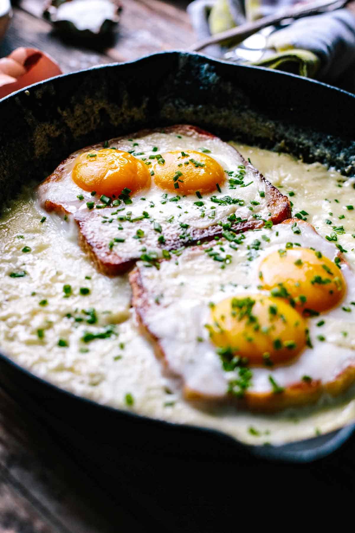 A close-up of baked eggs in a cast iron skillet showing two sunny-side up eggs with bright yellow yolks sitting atop ham or bacon, garnished with fresh chopped chives, and surrounded by a creamy sauce that's bubbling around the edges.