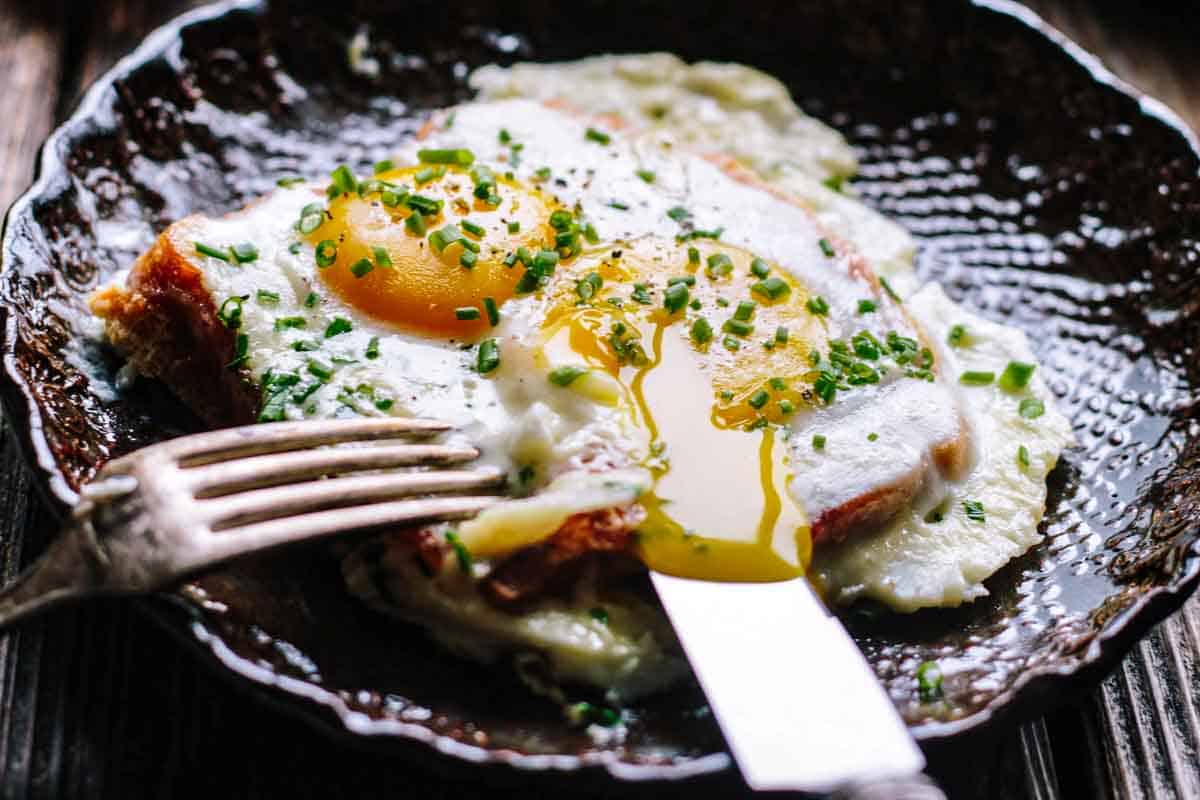 A rustic presentation of baked eggs in a dark, scalloped-edge pan on a wooden surface. One egg yolk has been broken with a fork, releasing golden yolk that mingles with the cream sauce. Silverware is positioned to the left, highlighting the dish's ready-to-eat appeal.