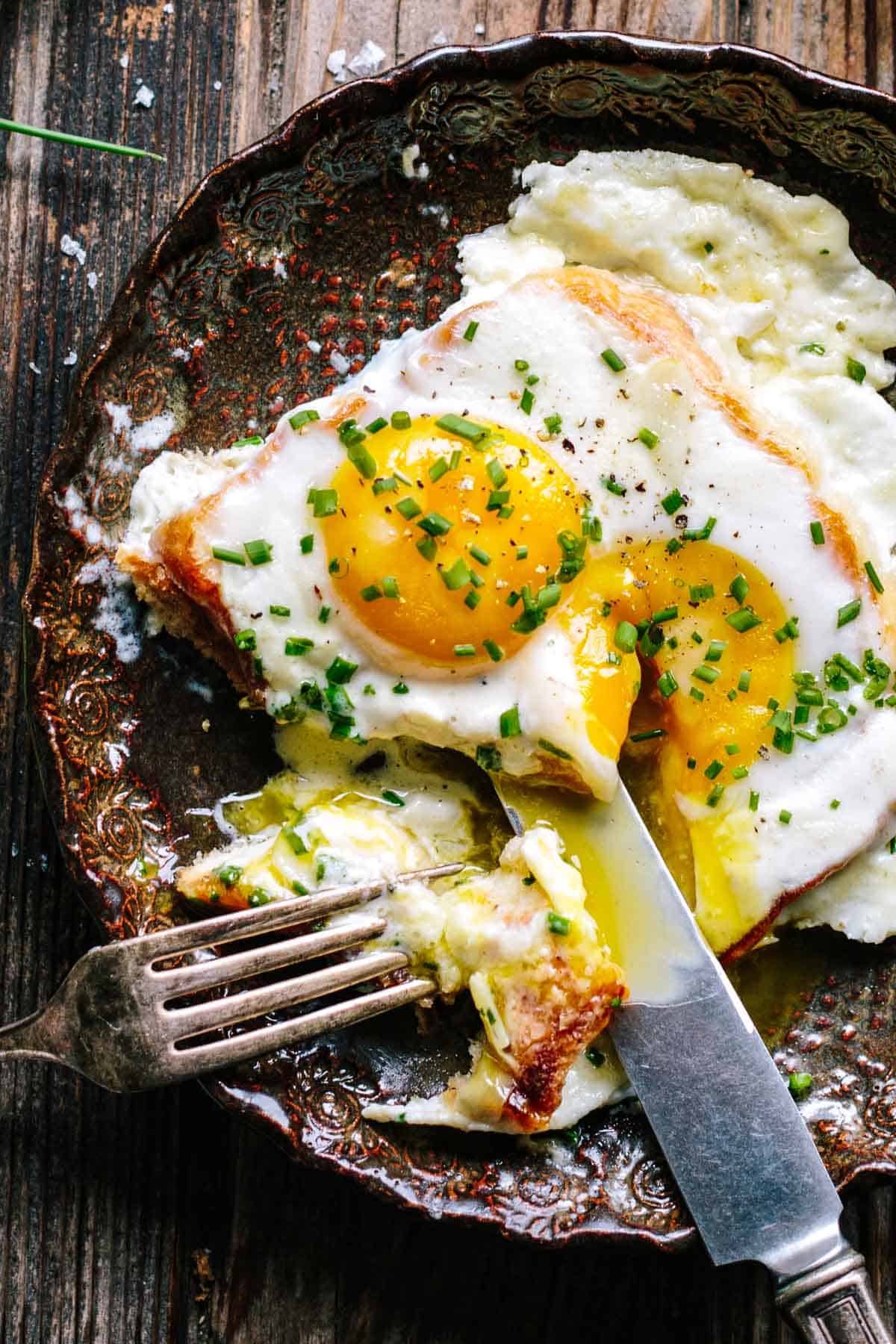 A decorative cast iron dish containing two sunny-side up eggs with vibrant yellow yolks atop what appears to be ham, sprinkled with chopped chives. A fork has pierced a portion, showing the creamy texture beneath. The dish sits on a weathered wooden surface.