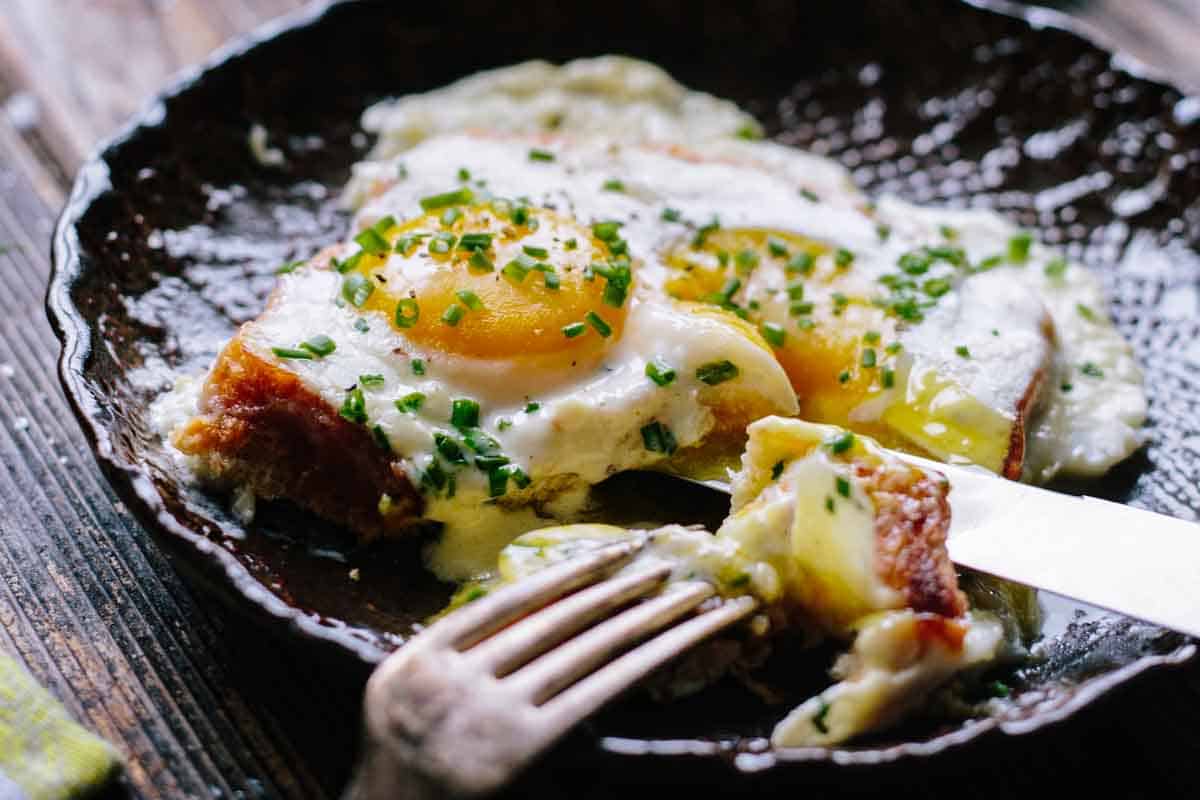 Another angle of the baked eggs in a decorative cast iron dish, showing the eggs nestled on ham with cream sauce. A bite has been taken with the fork visible in the frame, demonstrating the rich, creamy consistency of the dish served on rustic wooden boards.