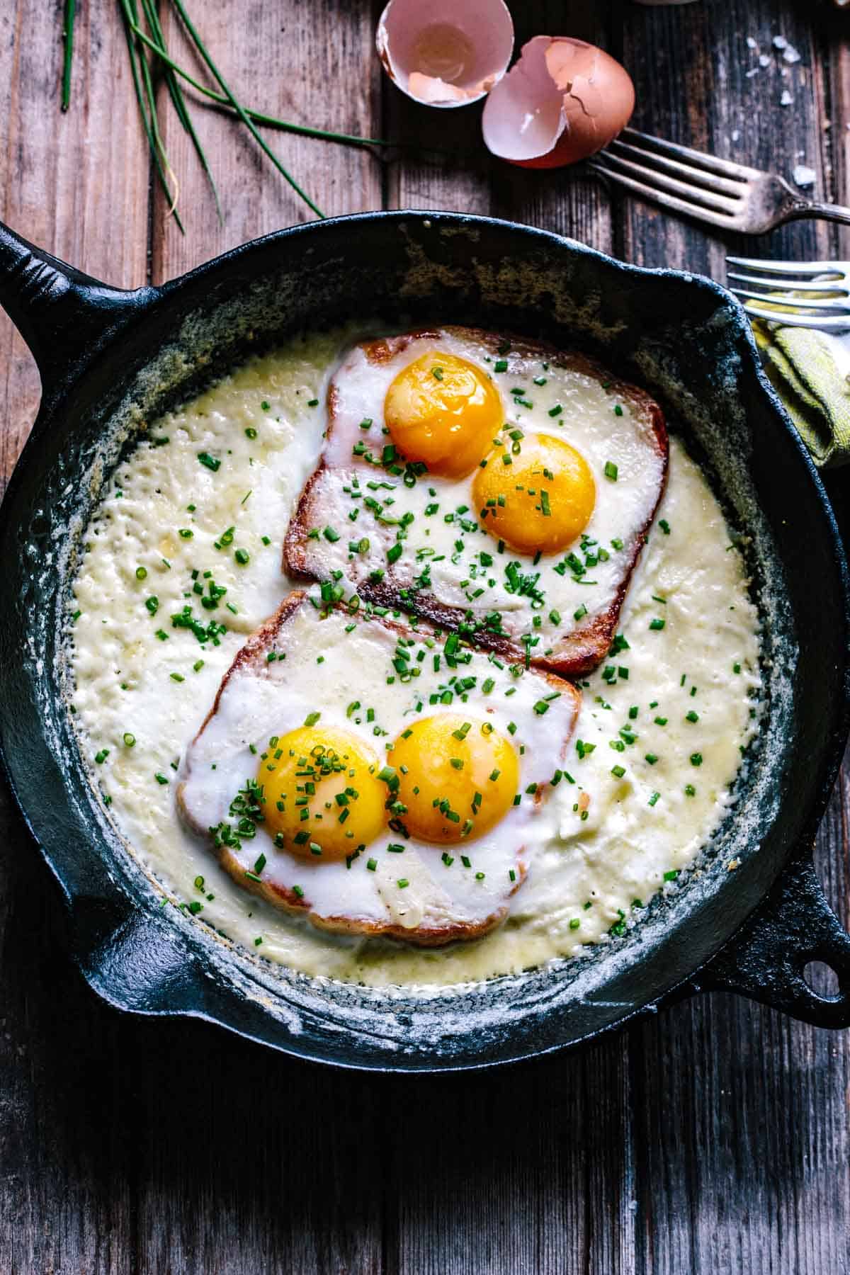 An overhead view of a cast iron skillet containing two slices of ham topped with four eggs (two on each slice), all baking in a creamy sauce and sprinkled generously with fresh chives. Cracked eggshells and fresh chives are visible in the background, emphasizing the dish's homemade quality.