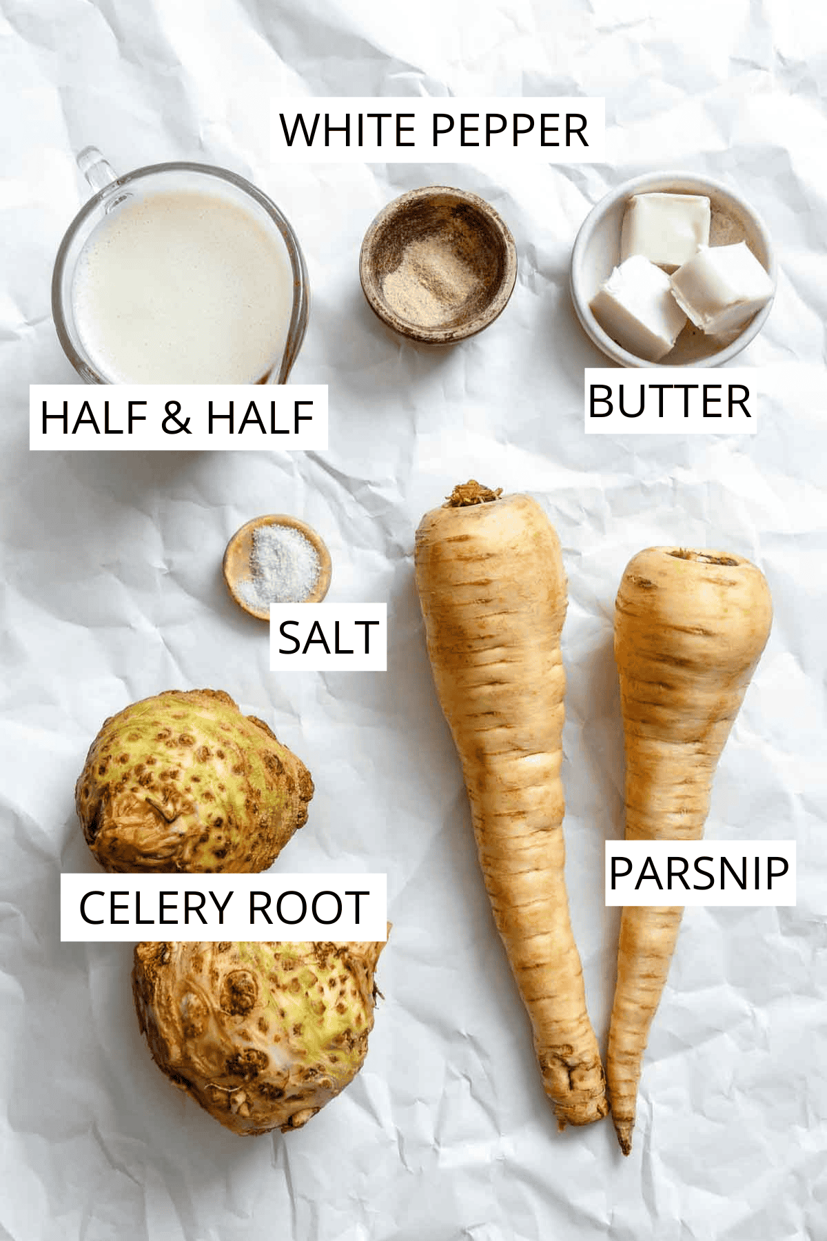 Ingredients for celery root and parsnip puree arranged on white parchment paper: two whole celery root bulbs, two parsnips, cubes of butter, a measuring cup of cream or milk, and small bowls of salt and spices.