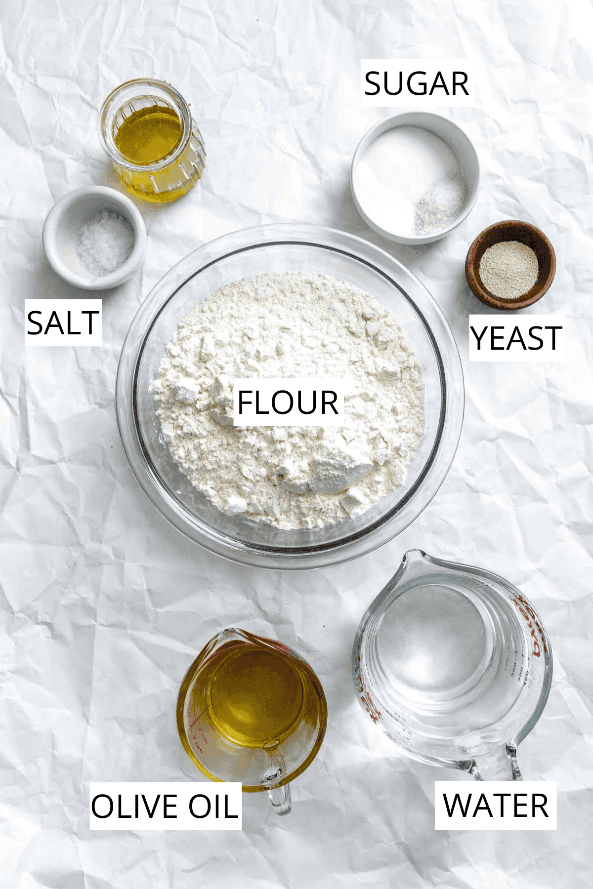 Overhead view of bread ingredients laid out on white paper including flour, olive oil, water, salt, and yeast.