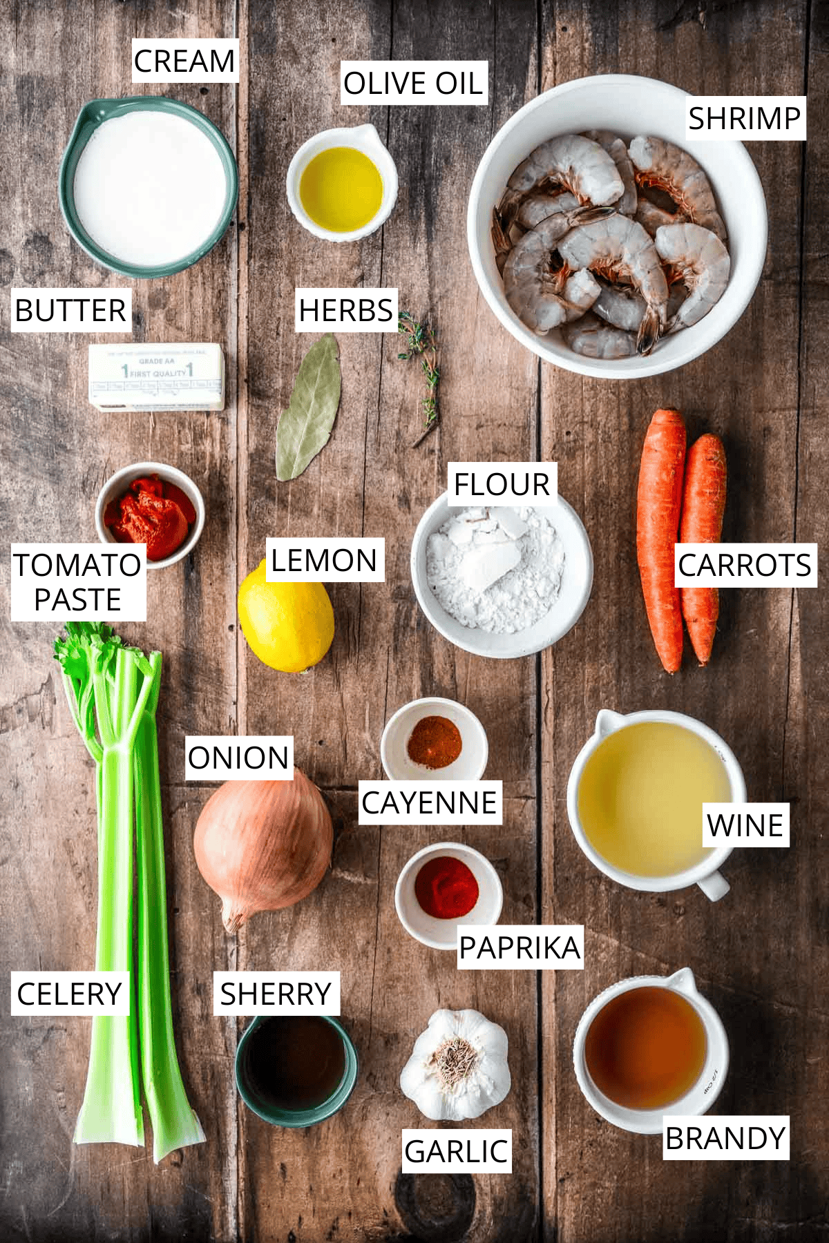 Overhead view of labeled ingredients for shrimp bisque arranged on a wooden surface, including cream, olive oil, butter, tomato paste, celery, onion, shrimp, herbs, flour, carrots, lemon, cayenne, paprika, garlic, sherry, wine, and brandy.