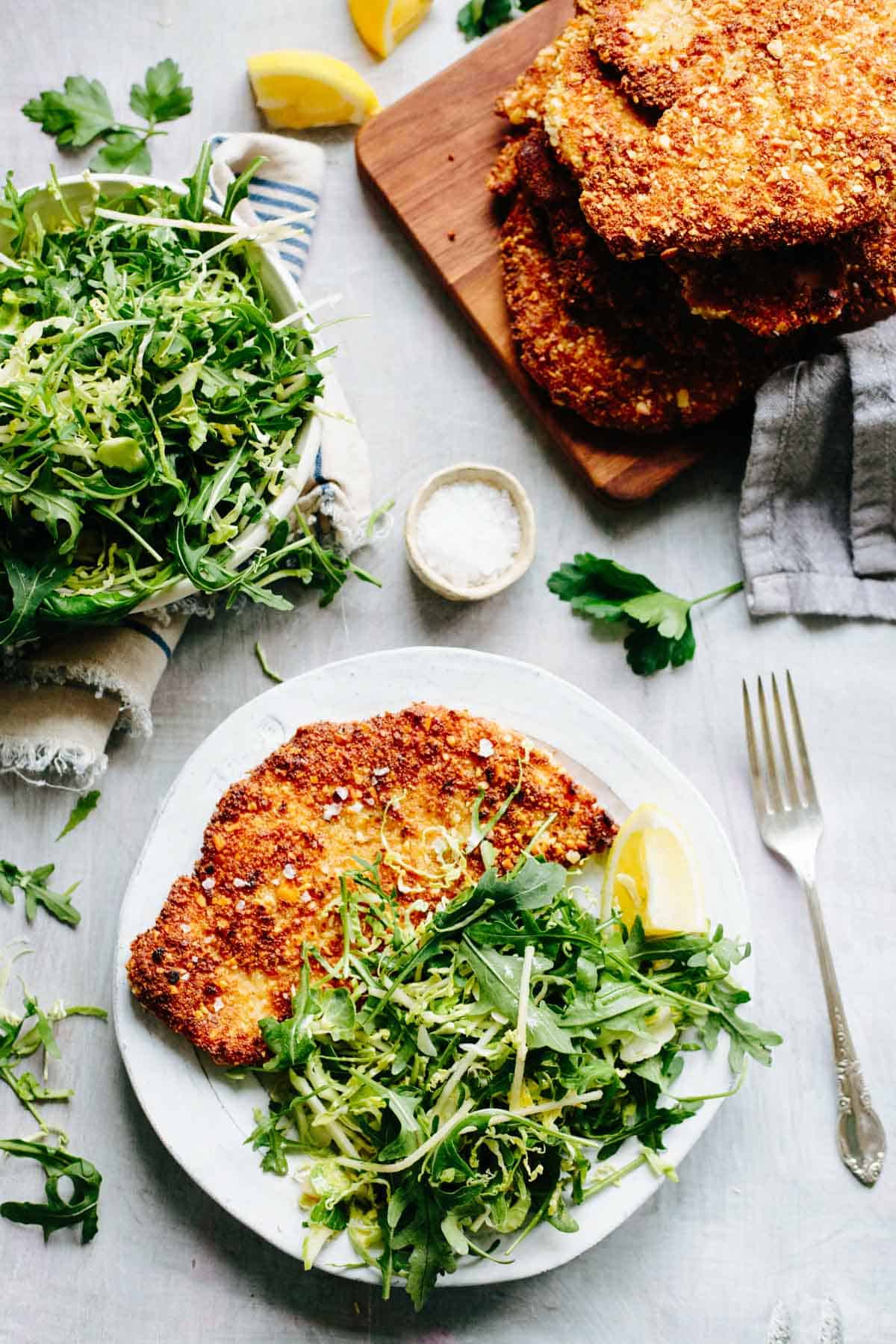 Stack of golden-brown almond crusted chicken cutlets on a wooden cutting board with lemon wedge, herbs, and kitchen towel.