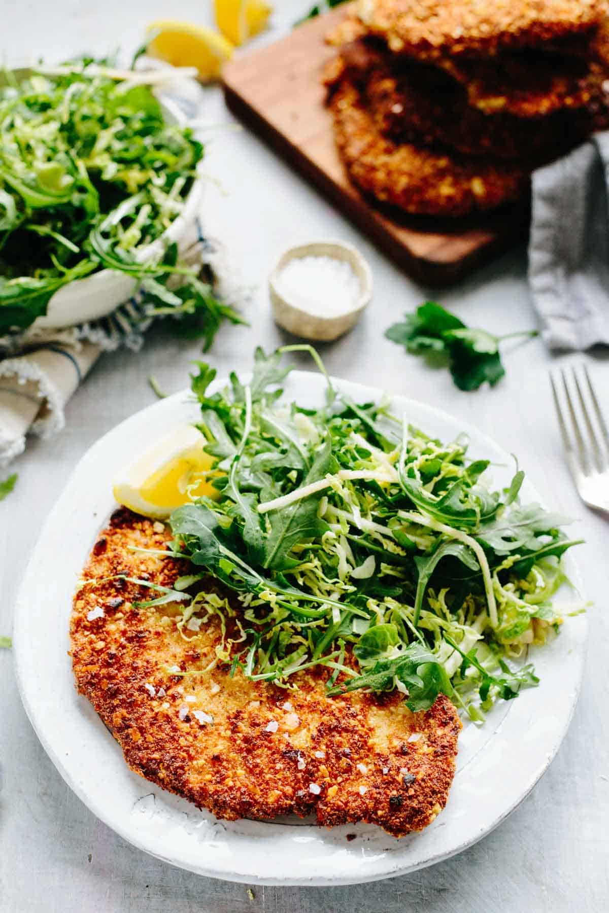 Overhead view of a plated almond crusted chicken cutlet with arugula salad, silverware, and additional elements of the meal visible.