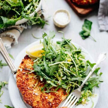 Close-up of almond crusted chicken cutlet on a white plate with vibrant green salad and lemon wedge, showing the golden crust and fresh garnishes.