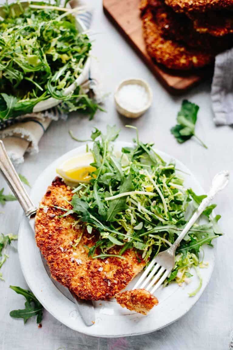 Close-up of almond crusted chicken cutlet on a white plate with vibrant green salad and lemon wedge, showing the golden crust and fresh garnishes.