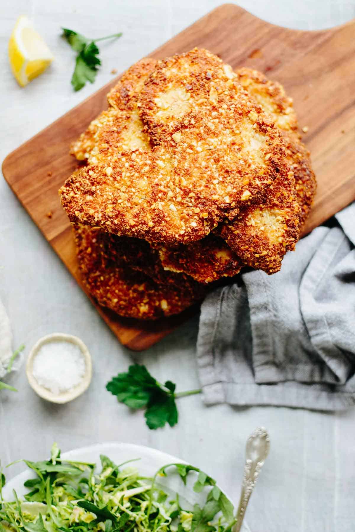 Overhead view of a plated almond crusted chicken cutlet served with fresh green salad and lemon wedge, with additional cutlets on a wooden board in the background.