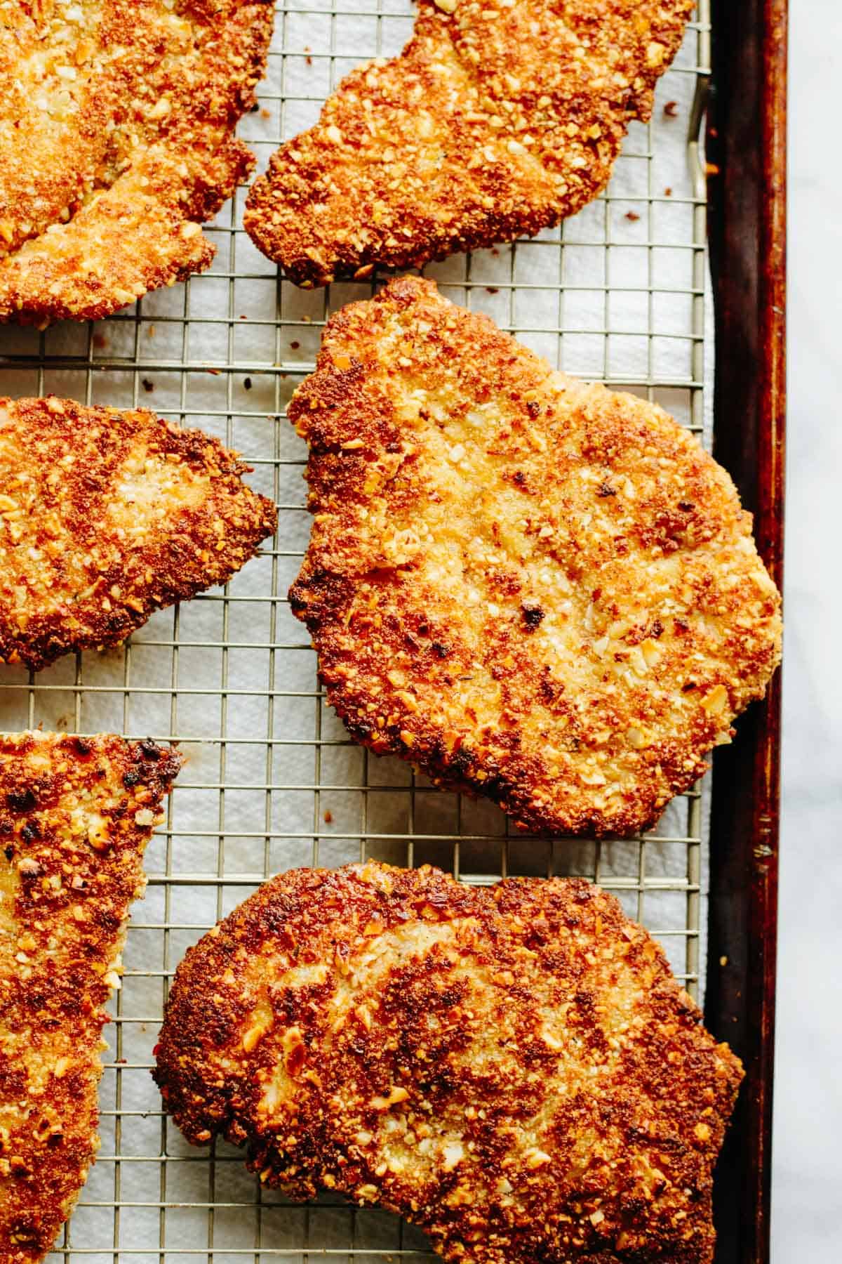 Golden-brown almond crusted chicken cutlets arranged on a wire cooling rack atop a baking sheet, showing their crispy textured coating.