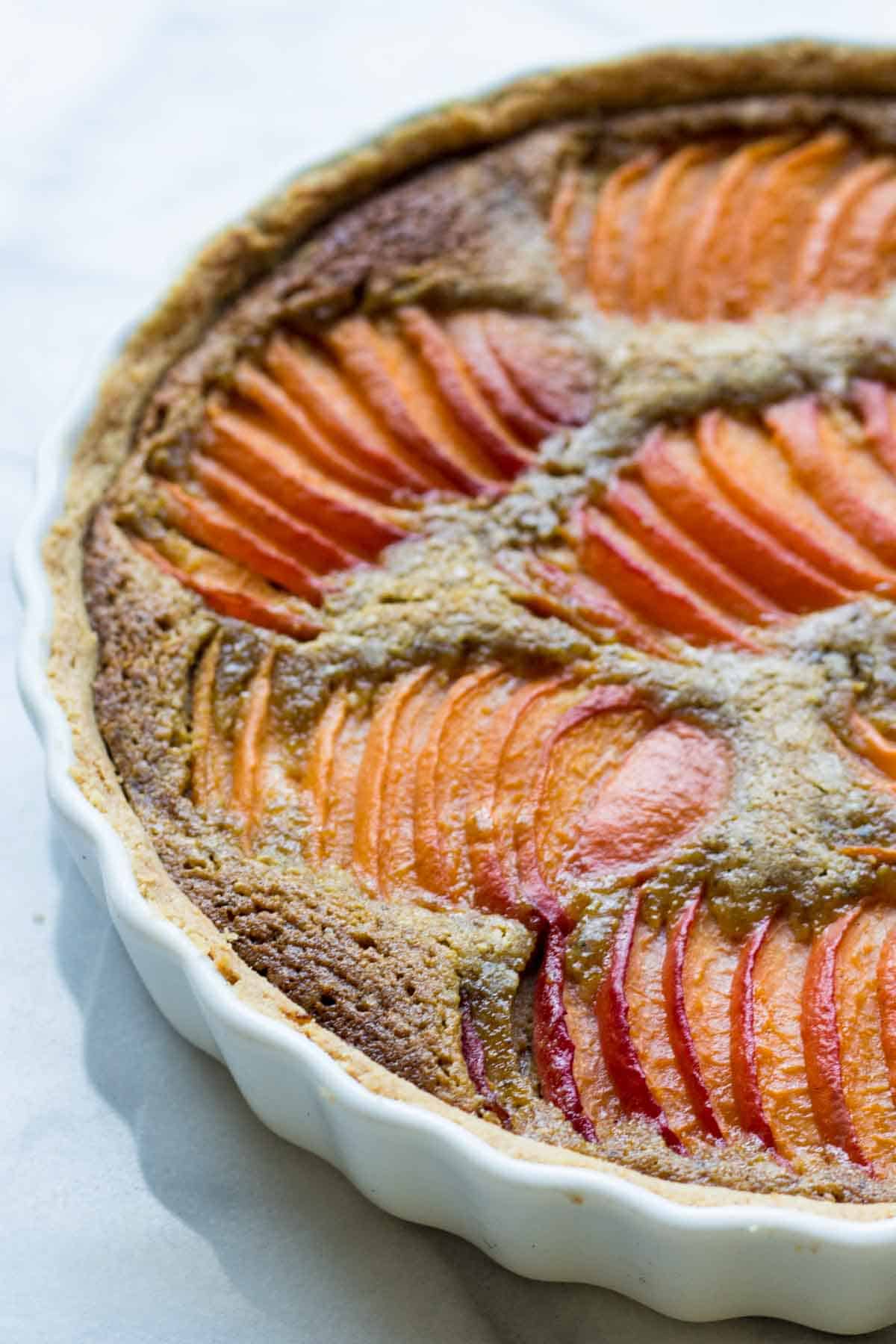 Close-up of a freshly baked apricot tart in a white ceramic dish, showing golden-orange apricot slices arranged in a pattern over a caramelized almond filling.