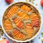 Overhead view of a complete apricot pistachio tart in a white ceramic dish, surrounded by fresh apricots and roasted pistachios on a marble surface.