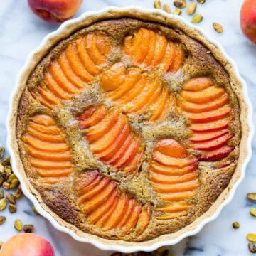 Overhead view of a complete apricot pistachio tart in a white ceramic dish, surrounded by fresh apricots and roasted pistachios on a marble surface.