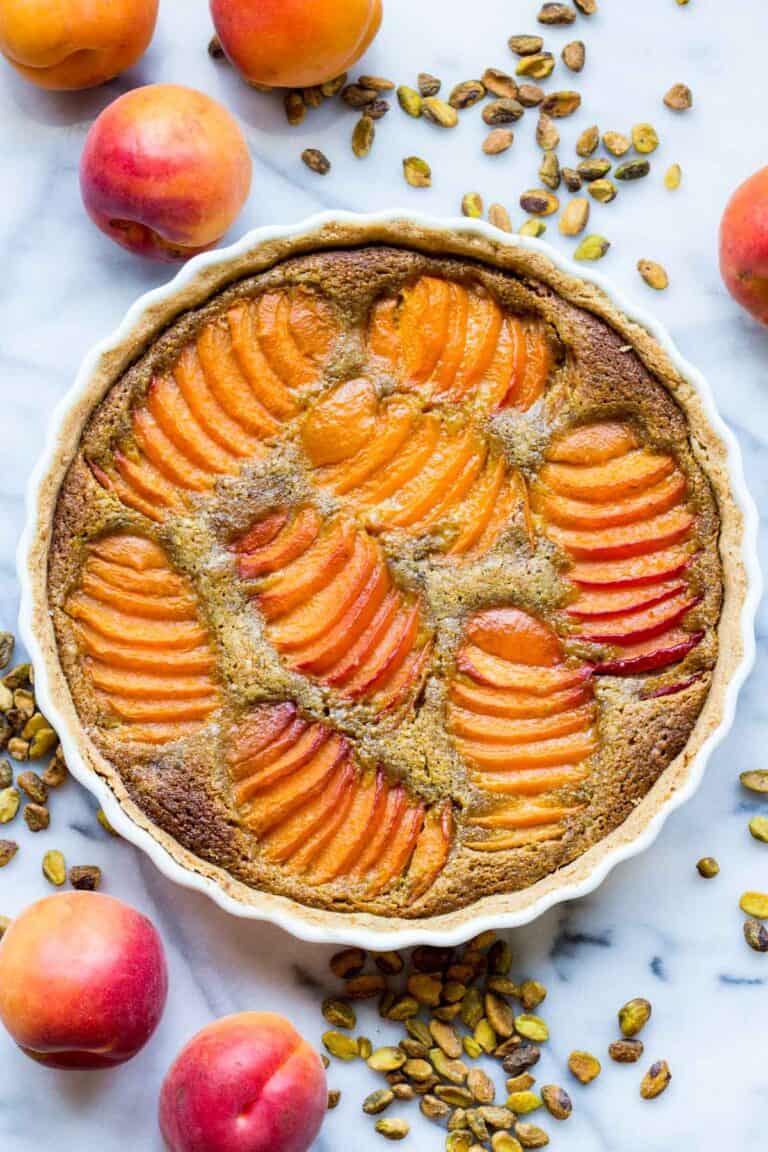 Overhead view of a complete apricot pistachio tart in a white ceramic dish, surrounded by fresh apricots and roasted pistachios on a marble surface.