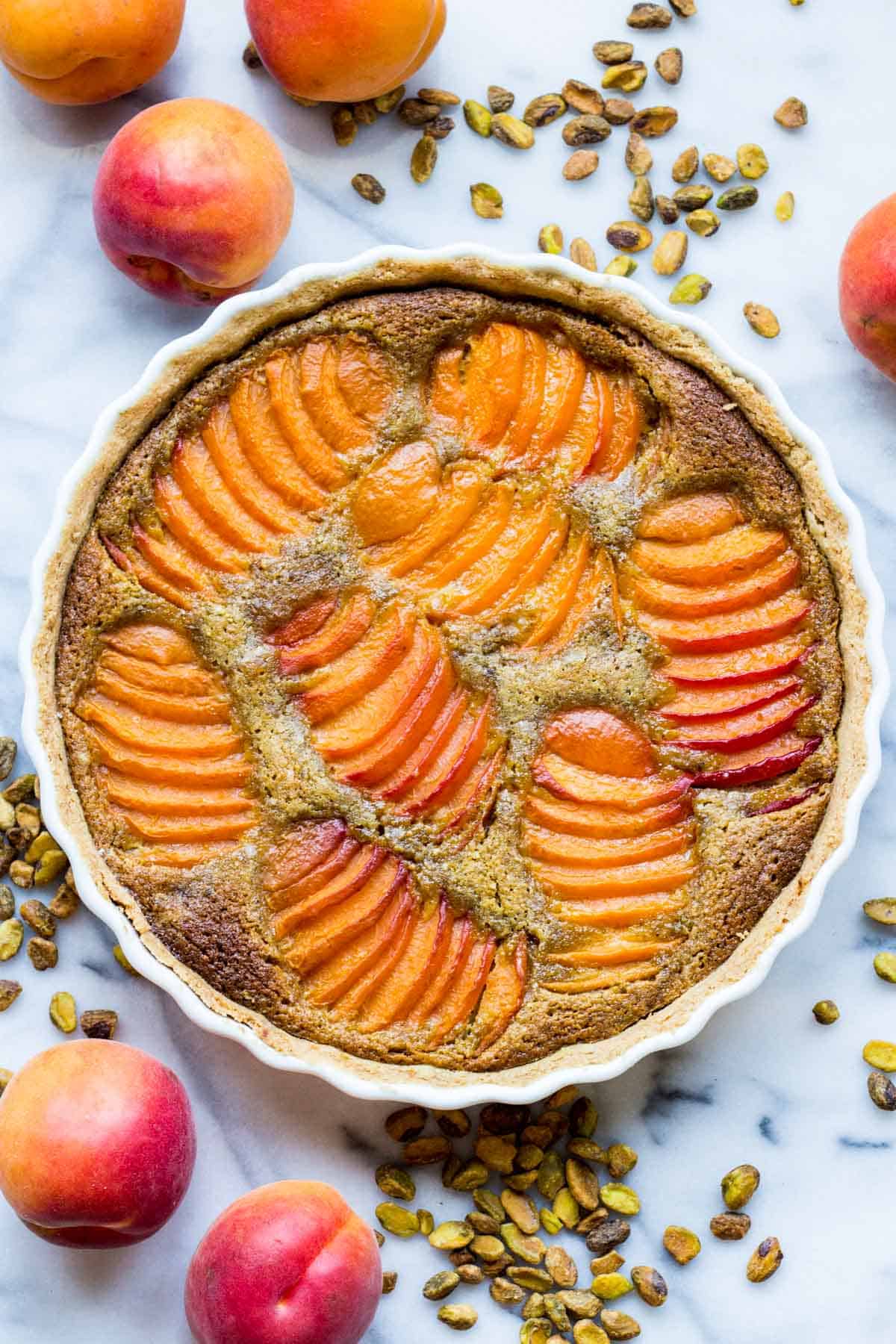 Overhead view of a complete apricot pistachio tart in a white ceramic dish, surrounded by fresh apricots and roasted pistachios on a marble surface.