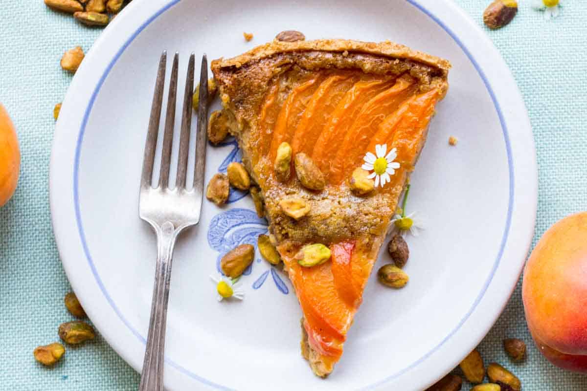 A slice of apricot pistachio tart on a blue-rimmed white plate, garnished with pistachios and a small daisy flower, with apricots visible in the background.