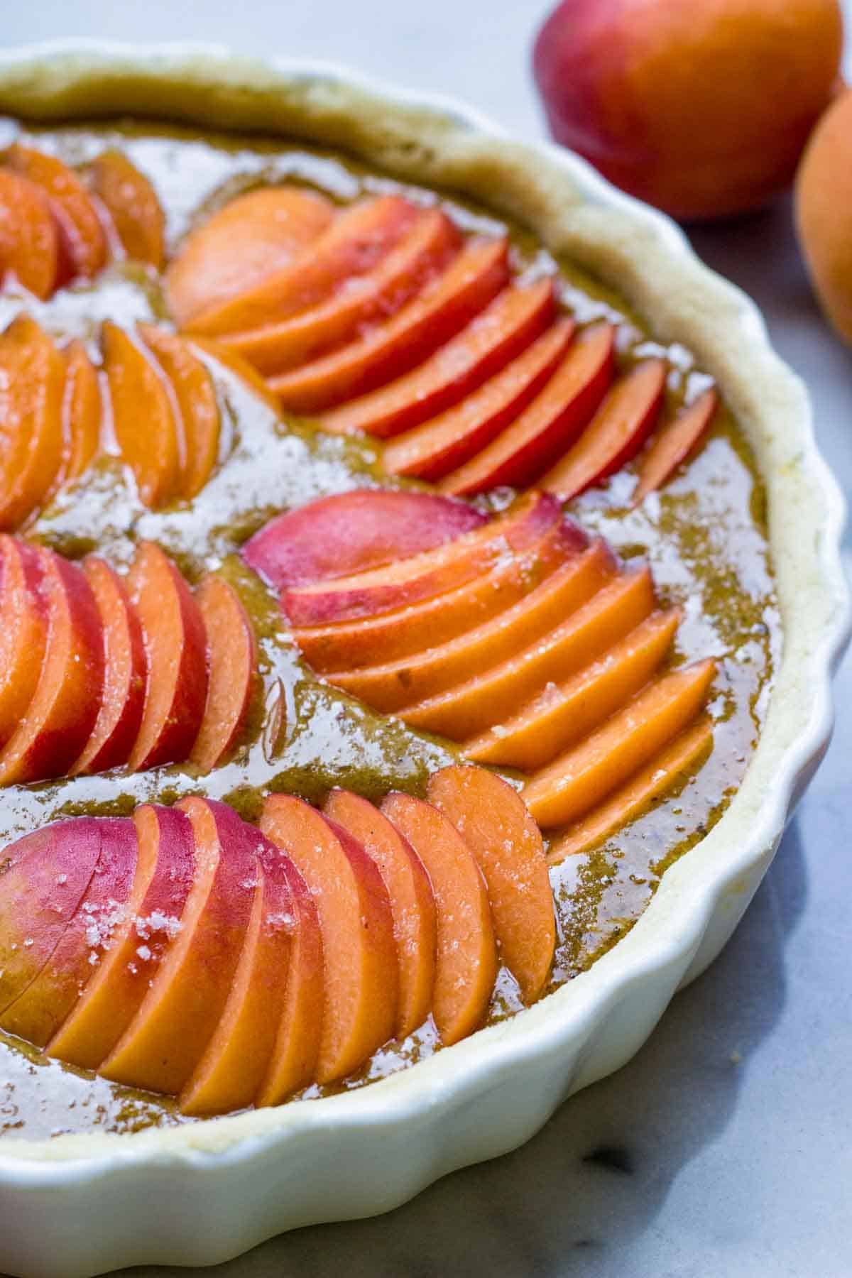 Close-up of an unbaked apricot tart showing thinly sliced fresh apricots arranged in a pattern and sprinkled with sugar in a white ceramic tart dish.