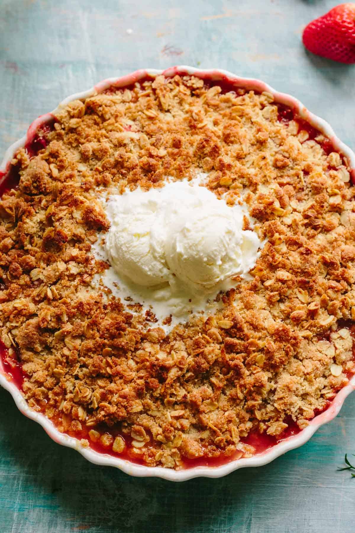 Freshly baked strawberry crisp in a white ceramic dish with a golden oat topping and vanilla ice cream in the center, served on a turquoise surface.