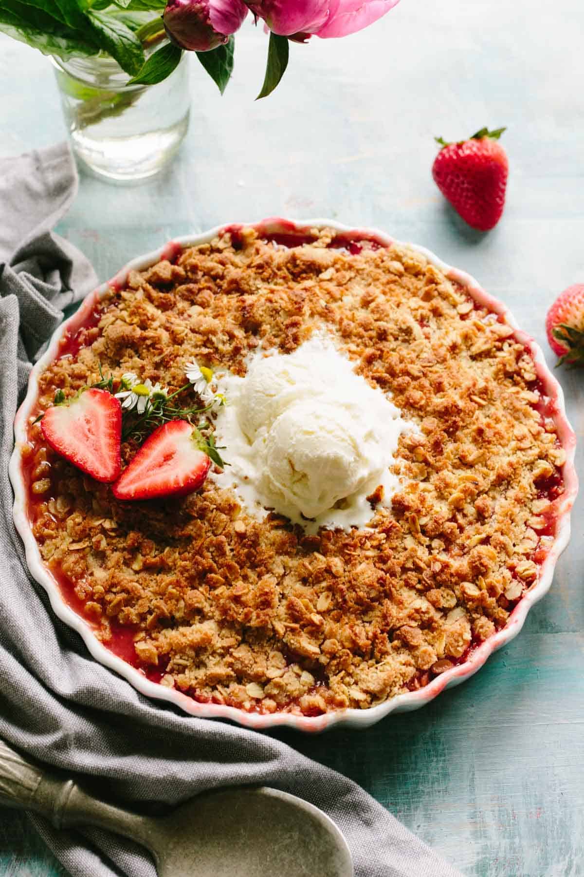 Overhead view of strawberry crisp in a white scalloped dish topped with vanilla ice cream, fresh strawberry slices, and small white flowers, with gray linen and strawberries in the background.