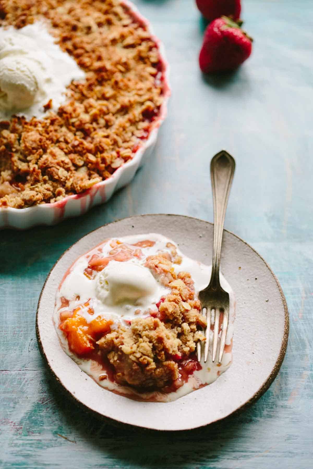 A serving of strawberry crisp on a white plate with melting vanilla ice cream and a silver fork, with the full crisp dish visible in the background.
