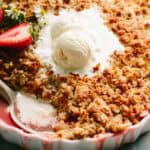 Close-up of strawberry crisp with a spoonful removed showing the bright red filling, topped with vanilla ice cream and fresh strawberry slices.