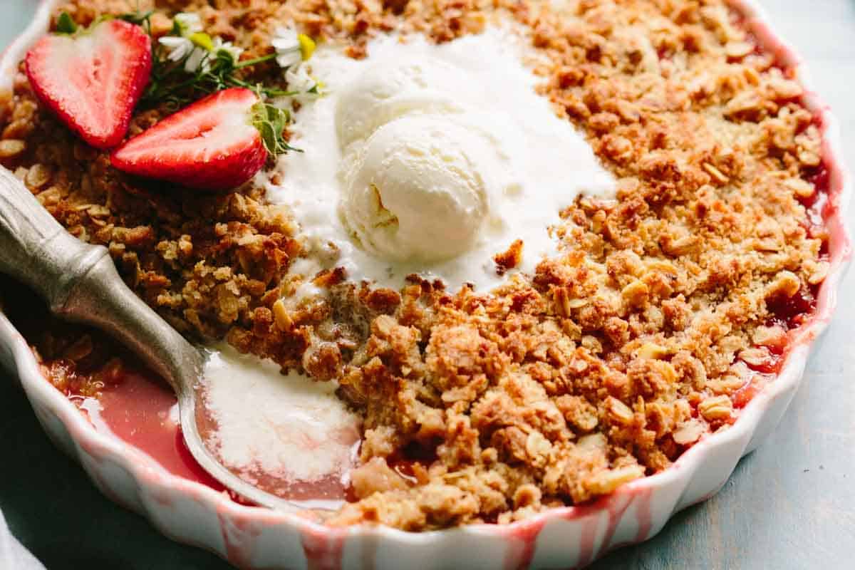 Close-up of strawberry crisp with a spoonful removed showing the bright red filling, topped with vanilla ice cream and fresh strawberry slices.