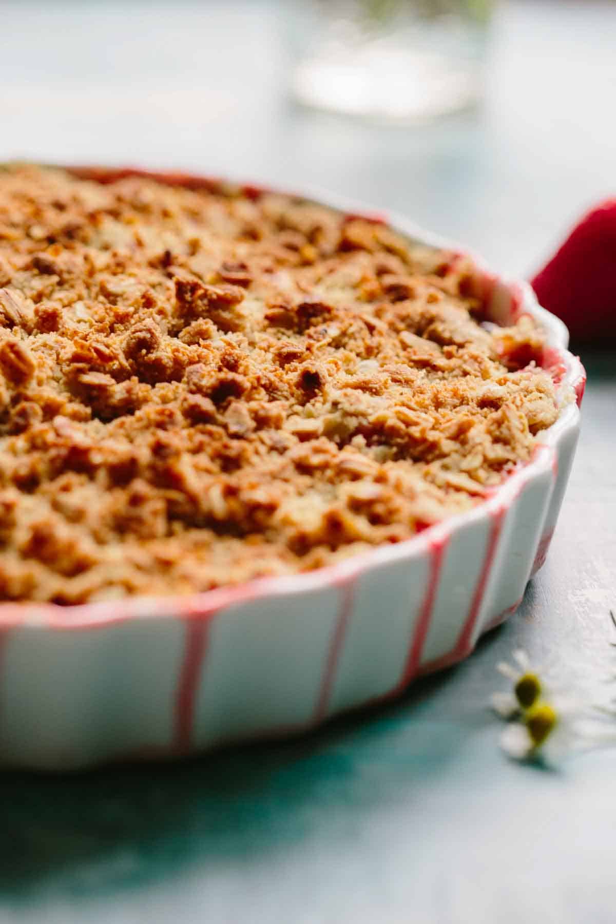 Side profile of a white ceramic dish filled with strawberry crisp showing the golden-brown oat topping, with a soft-focus background and hints of red.