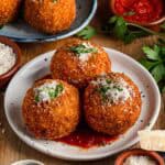 Table setting with white and blue plates containing arancini (stuffed rice balls) topped with grated cheese and herbs, served with marinara dipping sauce and garnishes.