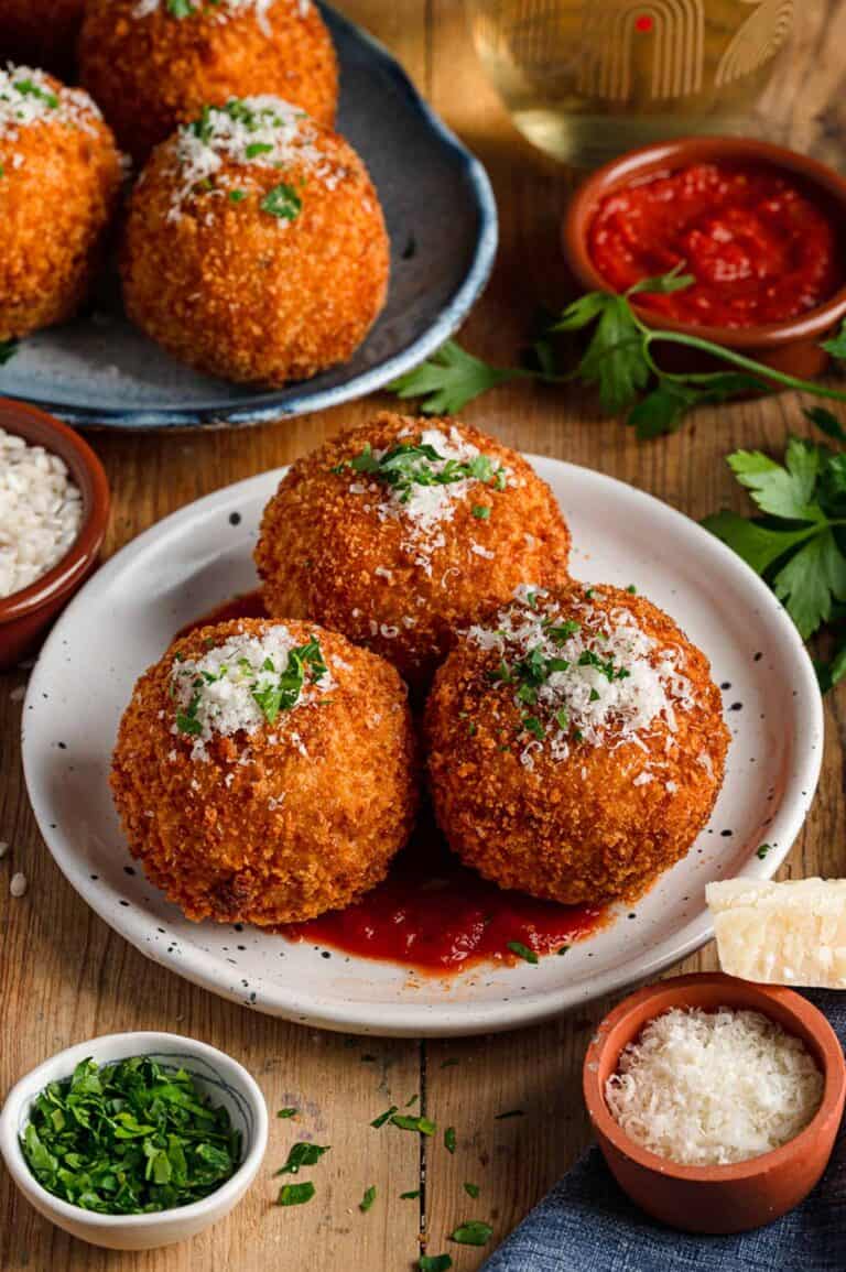 Table setting with white and blue plates containing arancini (stuffed rice balls) topped with grated cheese and herbs, served with marinara dipping sauce and garnishes.