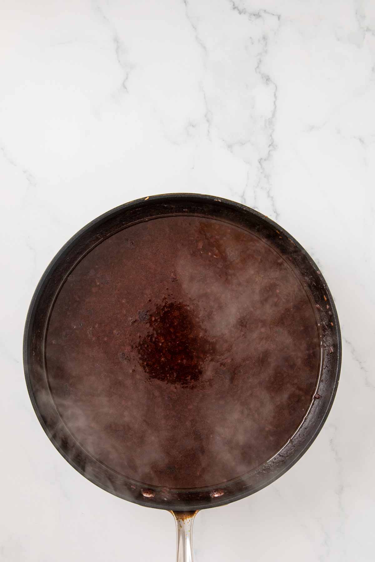 A black skillet on white marble containing a dark liquid, likely reduced red wine or the beginning stage of bordelaise sauce, with a reddish-brown color and glossy surface.