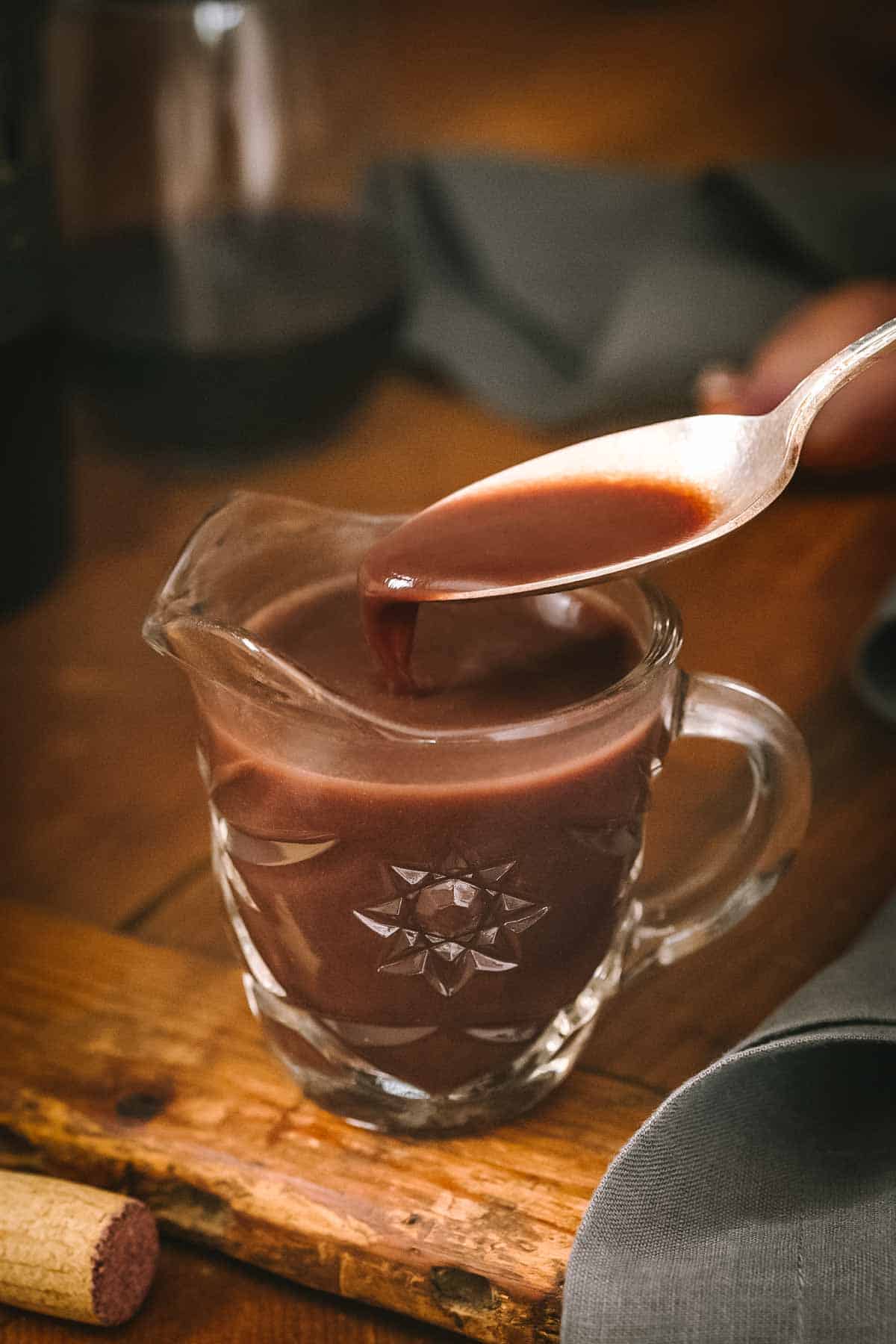 A glass measuring cup filled with rich brown bordelaise sauce, with a spoon drizzling some of the sauce back into the cup. The sauce has a thick, velvety consistency characteristic of a well-reduced sauce.