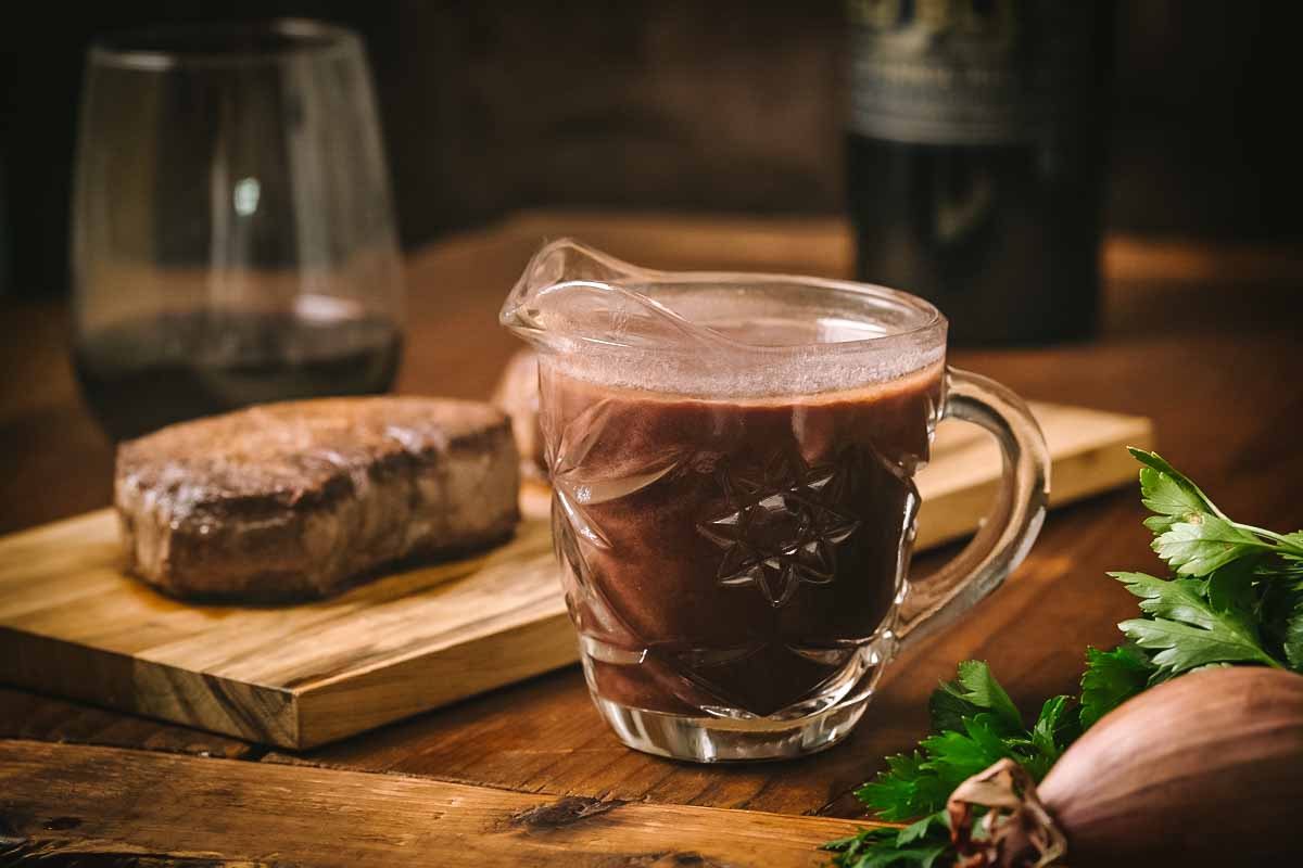 A glass pitcher of bordelaise sauce beside a grilled steak on a wooden cutting board. The sauce appears dark and glossy, ready to be served, with fresh herbs visible in the corner.
