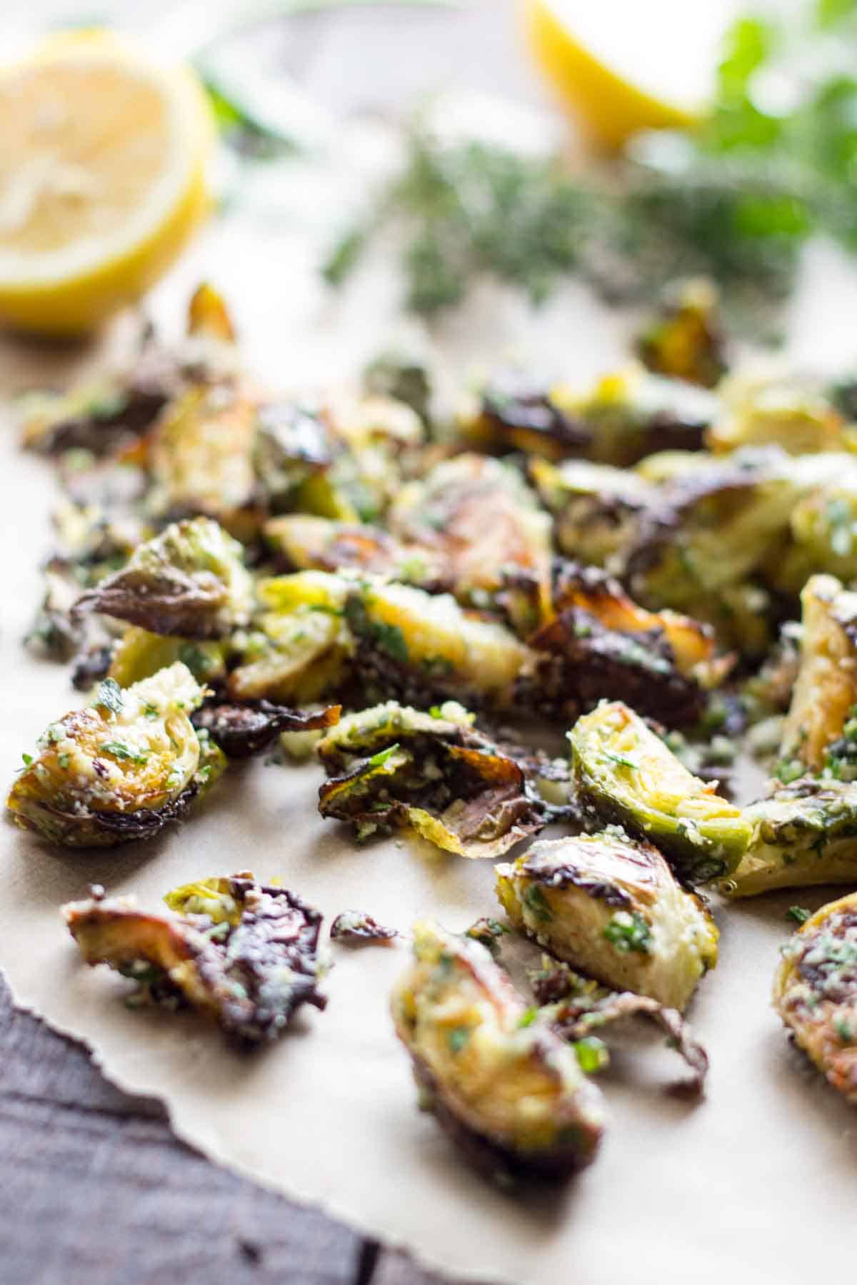 Roasted Brussels sprouts with crispy, caramelized edges scattered on parchment paper. The sprouts appear seasoned with herbs and grated cheese, with a lemon half visible in the blurred background, suggesting a bright citrus element to the dish.