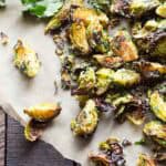 An overhead shot of roasted Brussels sprouts on parchment paper with fresh herbs and lemon visible in the frame. Fresh thyme sprigs and parsley leaves accompany the caramelized Brussels sprouts, which show varying degrees of browning and charring on their leaves.
