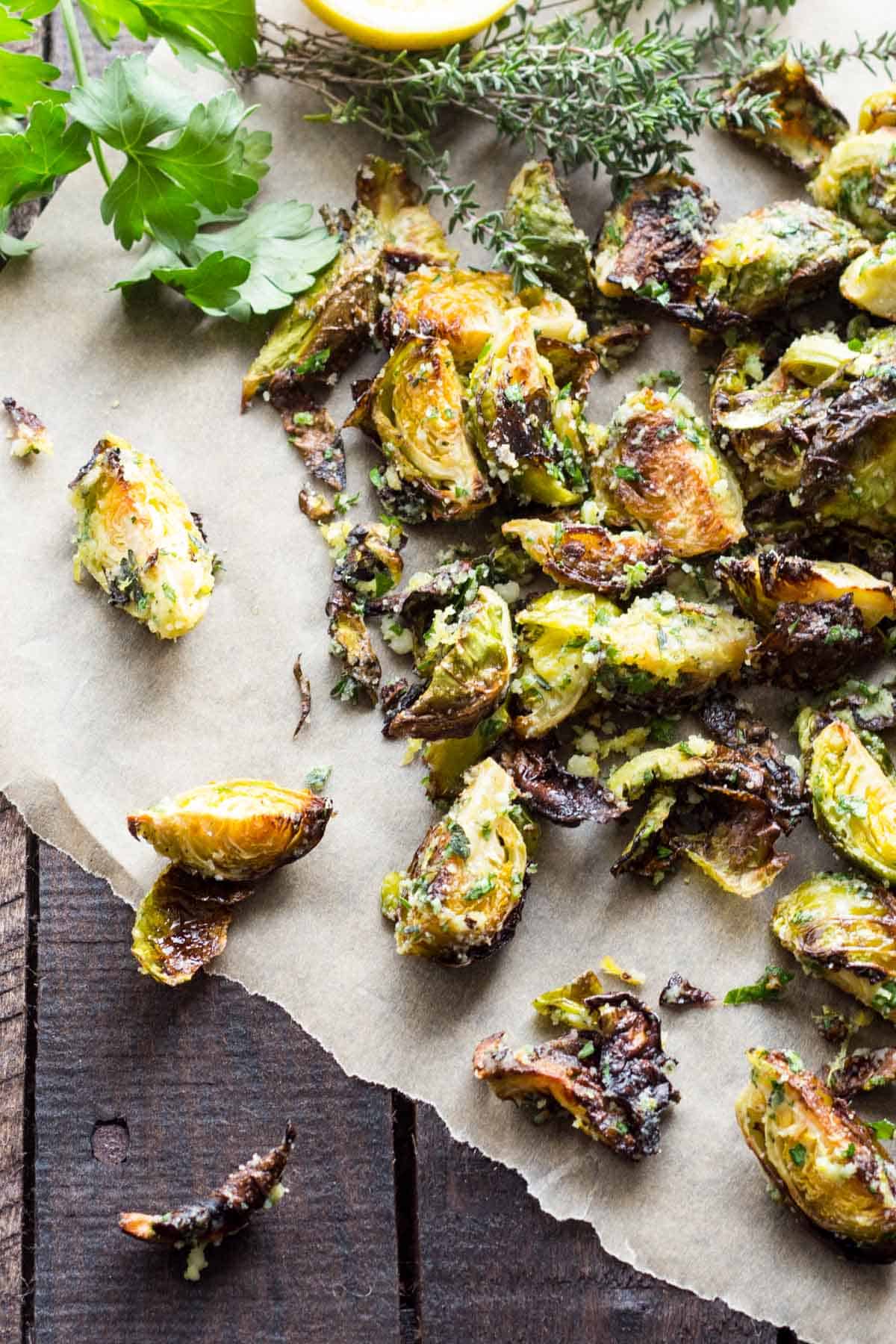 An overhead shot of roasted Brussels sprouts on parchment paper with fresh herbs and lemon visible in the frame. Fresh thyme sprigs and parsley leaves accompany the caramelized Brussels sprouts, which show varying degrees of browning and charring on their leaves.