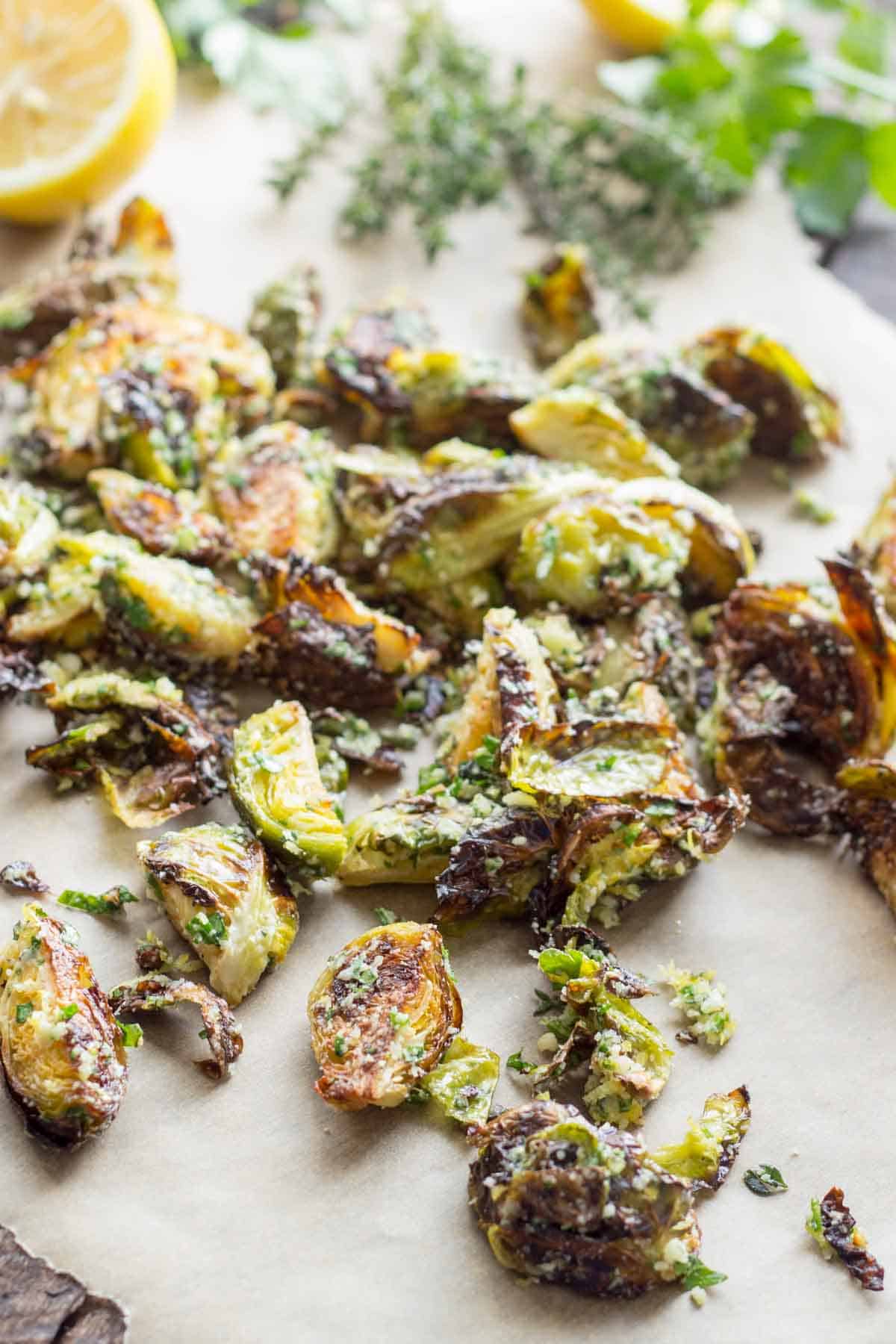 Another angle of the roasted Brussels sprouts on parchment paper, highlighting their crispy texture and the herb-cheese mixture coating them. The background shows fresh herbs and lemon, with the Brussels sprouts displaying a beautiful contrast between their charred exterior and bright green interior.