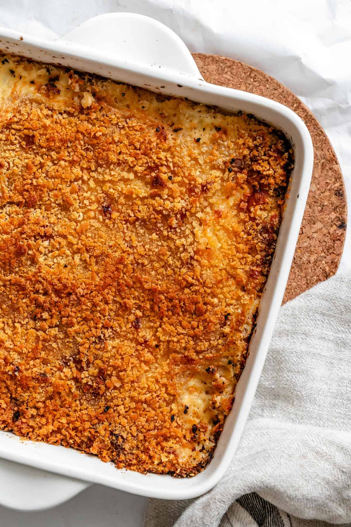 A close-up of the golden-brown, crispy-topped parsnip and celery root gratin fresh from the oven, showing the bubbly, browned breadcrumb crust in a white ceramic baking dish resting on a cork trivet.