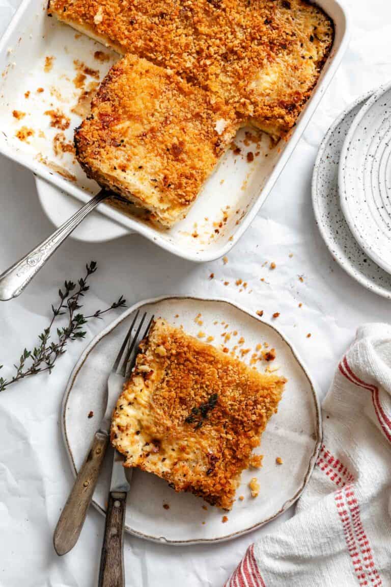 A serving of parsnip and celery root gratin on a white plate with a serving in the baking dish, showing the creamy, layered texture inside and the crispy top, accompanied by vintage utensils and a sprig of thyme.
