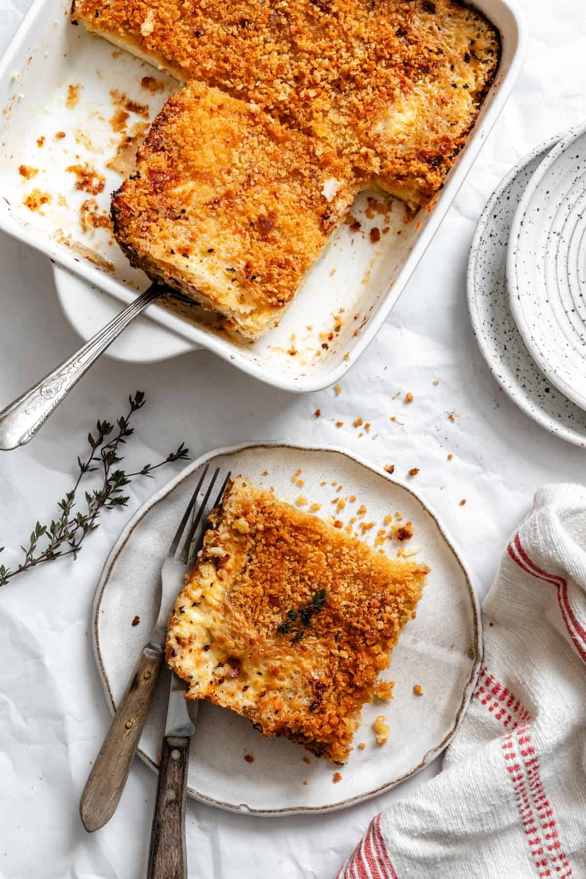 A serving of parsnip and celery root gratin on a white plate with a serving in the baking dish, showing the creamy, layered texture inside and the crispy top, accompanied by vintage utensils and a sprig of thyme.