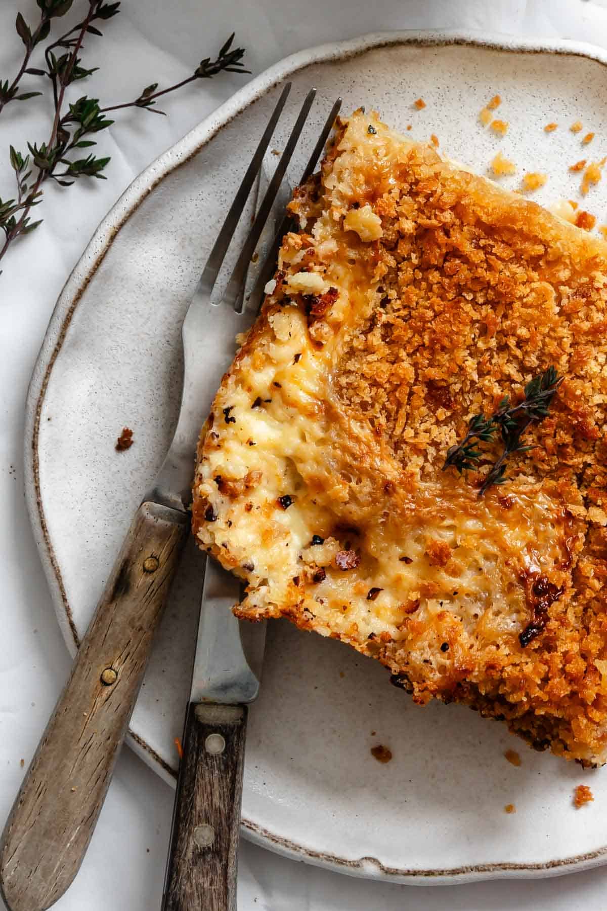 A close-up of a slice of parsnip and celery root gratin on a white ceramic plate, highlighting the layers of vegetables bound with creamy sauce, topped with the golden-brown crust and garnished with fresh thyme.