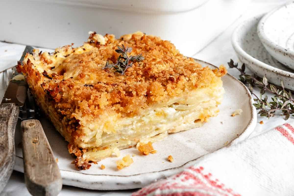 A side view of a thick slice of parsnip and celery root gratin on a white plate showing the distinct creamy layers of the root vegetables, with a crispy breadcrumb topping and vintage cutlery alongside.