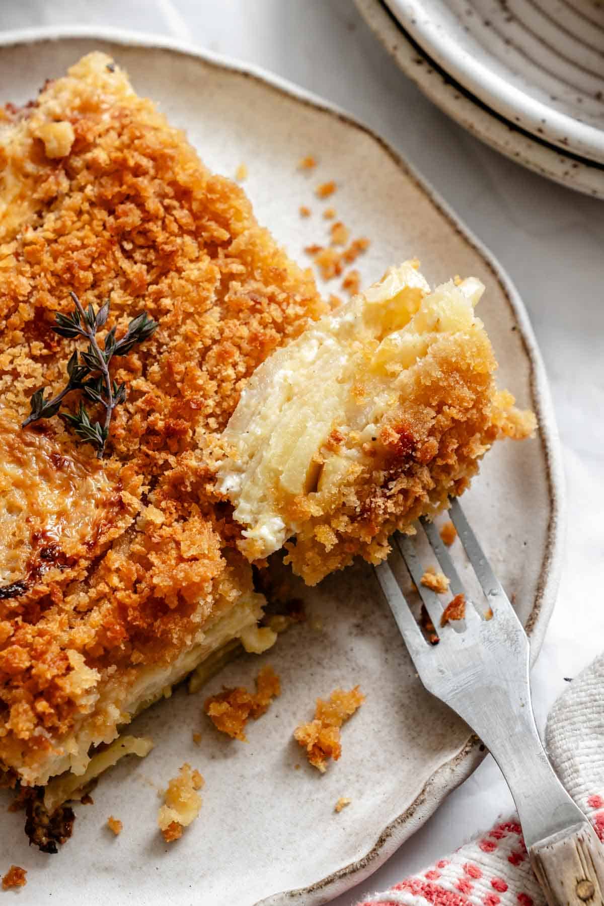 An extreme close-up of a forkful of the parsnip and celery root gratin, showcasing the tender vegetable layers and creamy sauce, with the remaining portion visible on a rustic ceramic plate.