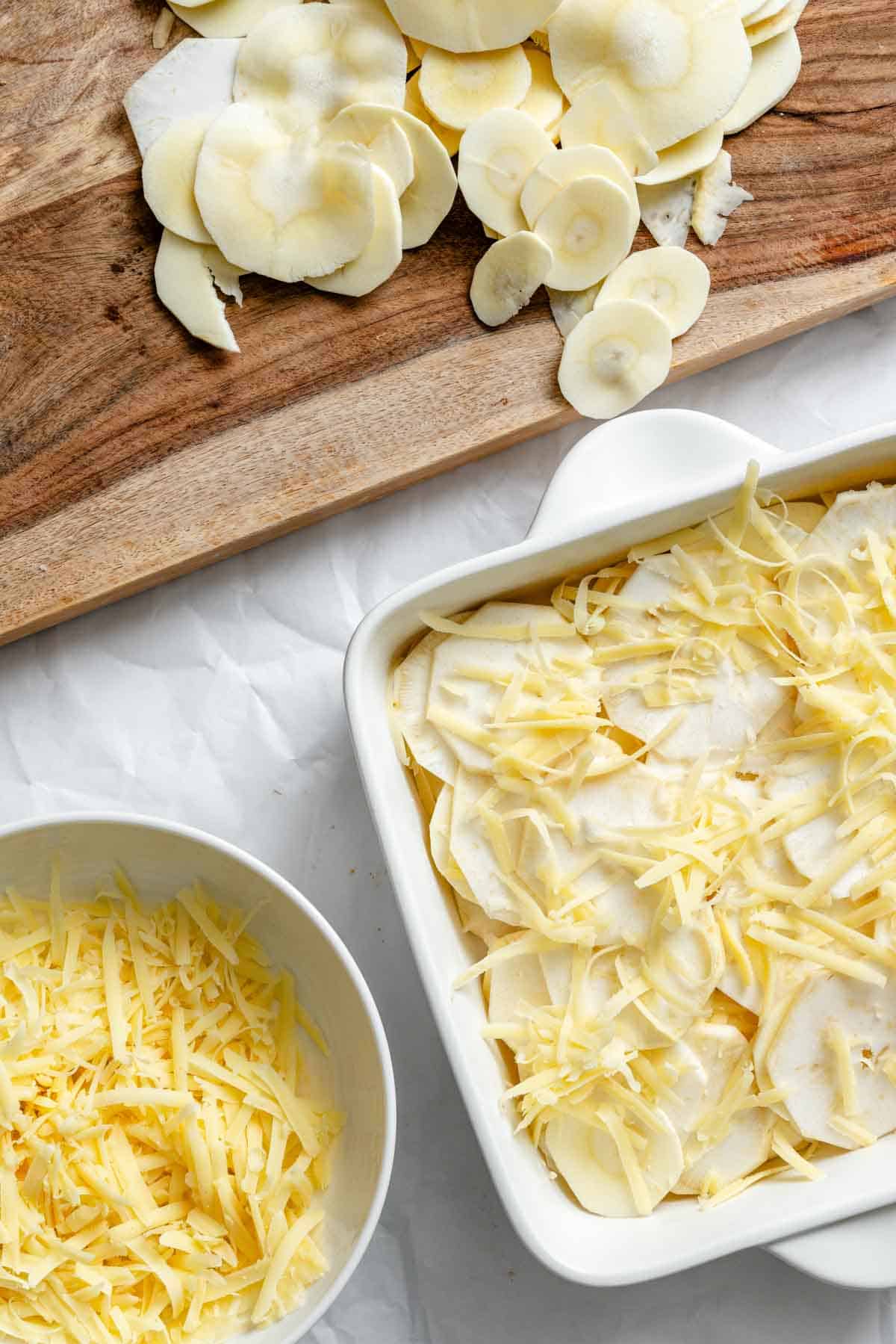 Thinly sliced white parsnips on a wooden cutting board with shredded cheese in a white bowl and a white baking dish filled with layered vegetables and cheese visible in the background, all set against a white parchment paper surface.